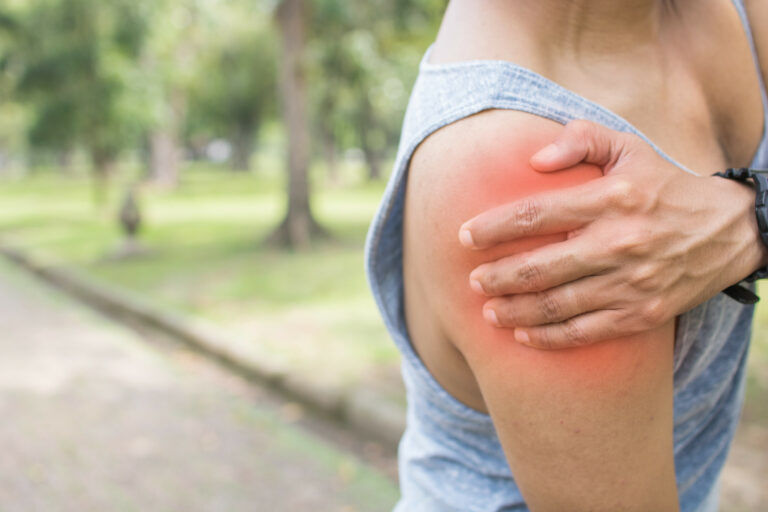 This is an image of someone jogging through a park who has stopped to grasp at pain in their shoulder, something that can at times be treated with Physical Therapy in Lewis Center by Athletic Advantage.