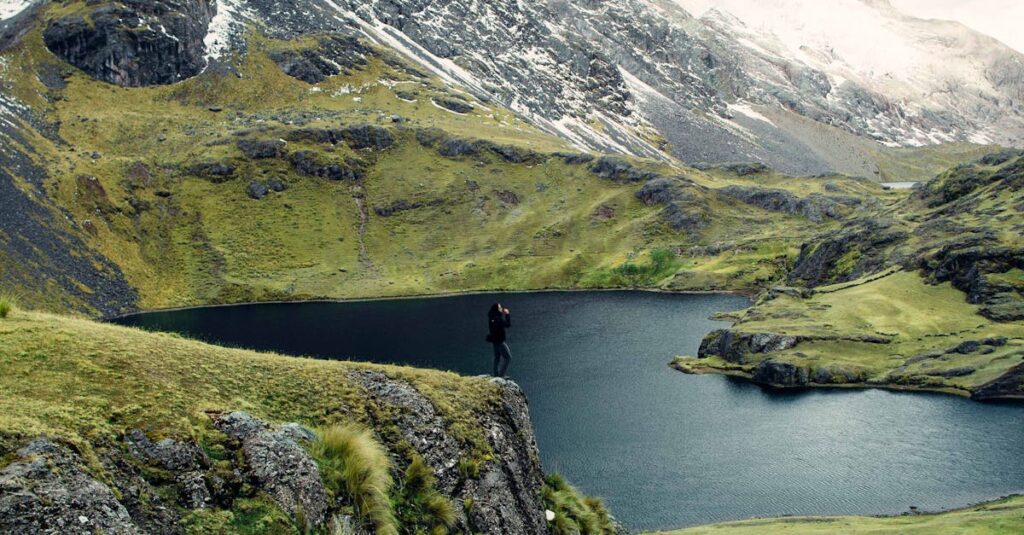 A hiker stands on a cliff overlooking a serene mountain lake surrounded by lush greenery and snow-capped peaks.
